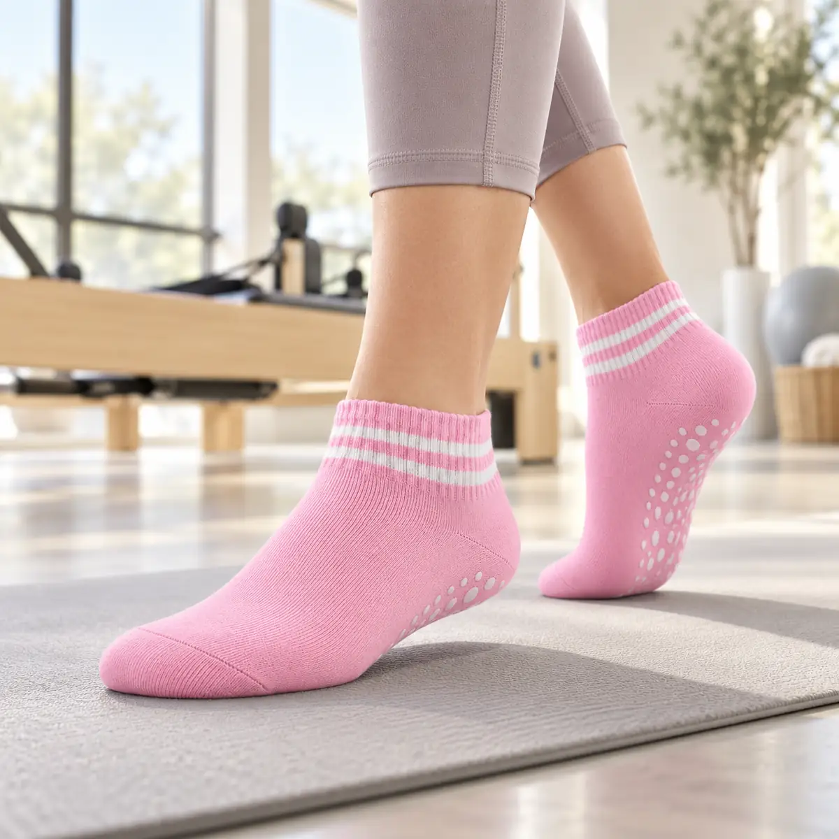 Bild 1 von 3 Rosa Knöchel-Socke von Trittfein mit weißen Streifen auf einer grauen Matte, Noppen unter der angehobenen Ferse, helles Studio mit Reformer unscharf im Hintergrund